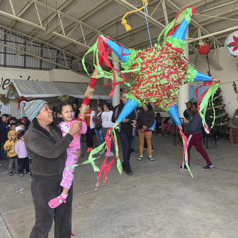 In this photo provided by Father Brian Strassburger, a mother holds her daughter as they swing at a piñata as other migrants at the shelter line up at the Casa del Migrante shelter in Reynosa, Mexico, on Thursday, Dec. 22, 2022. Tens of thousands of migrants who fled violence and poverty will spend Christmas in crowded shelters or on the dangerous streets of Mexican border towns. (Father Brian Strassburger via AP)