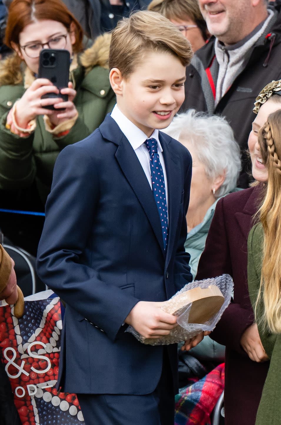 prince george of wales smiles as he stands next to people, he holds a wooden plaque in his hands and wears a blue suit with a white collared shirt and blue patterned tie