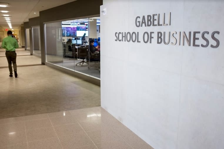 Interior hallway with Gabelli School of Business sign on wall to the right. Beyond sign there is a wall made of windows that has computers on the other side. The back of a man walking down the hallway can be seen in the background.