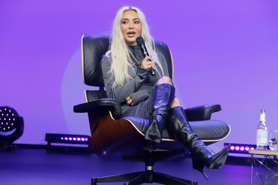 Kim Kardashian, seated on a modern chair, speaks into a microphone at an event. She is wearing a long-sleeve dress and knee-high boots