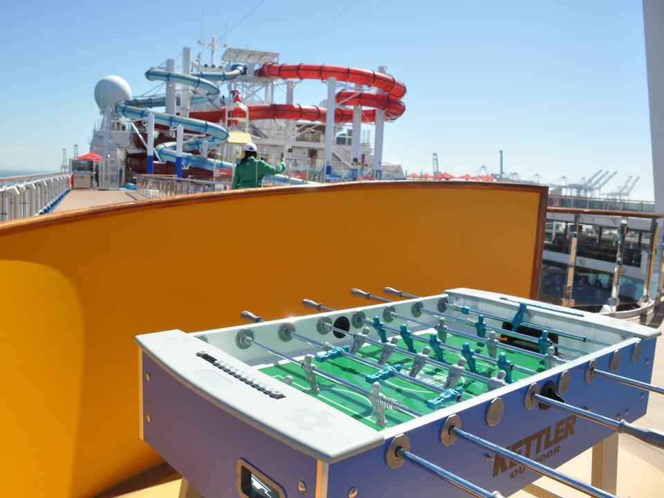 A foosball table on the top deck of a cruise ship with waterslides in the background.