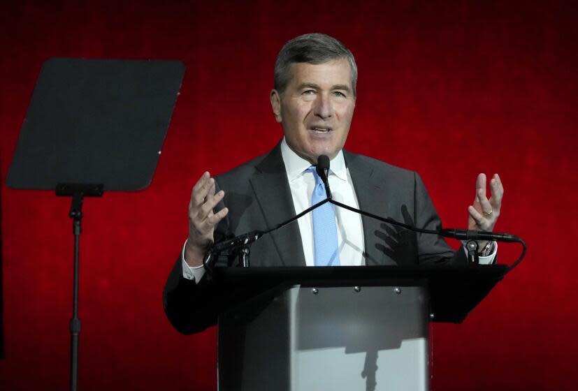 Charles Rivkin gestures at a podium while speaking into a microphone and wearing a gray suit with a blue tie.