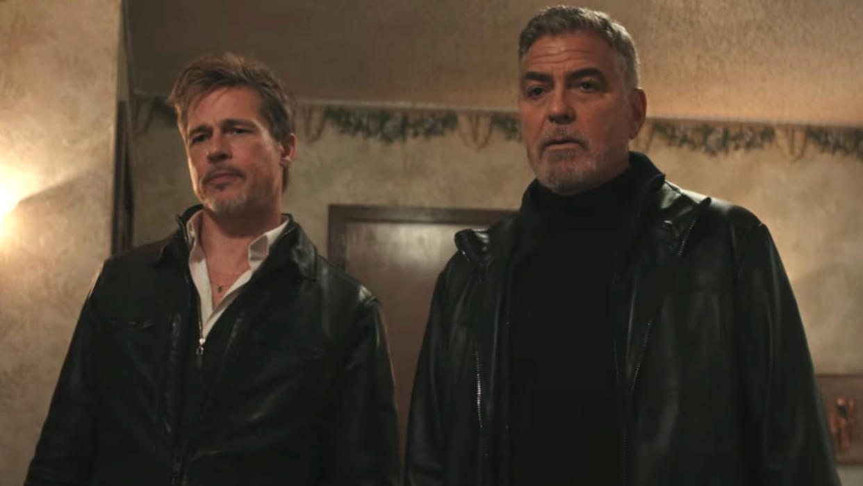  Brad Pitt and George Clooney standing together with looks of disbelief in Wolfs. 