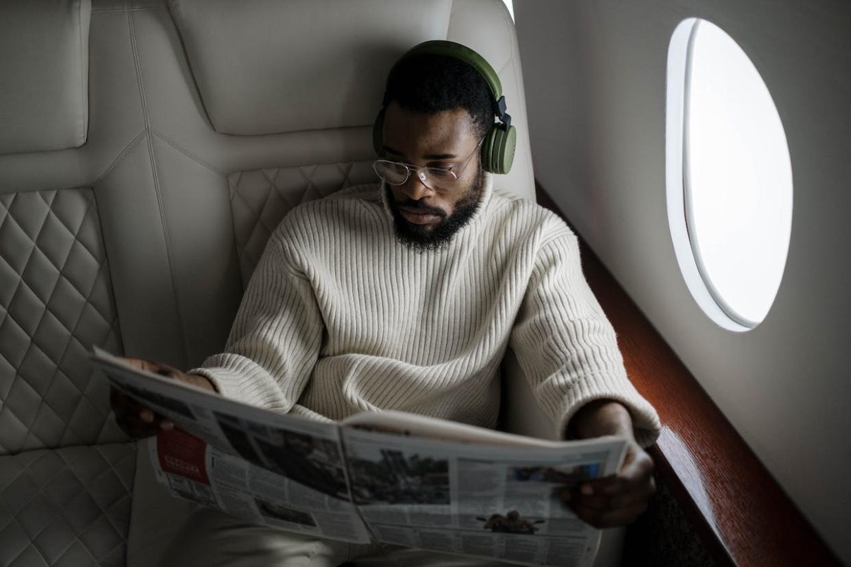Can you Use Bluetooth on a Plane and what happens if you do? Learn here. Pictured: a man wearing headphones while reading a newspaper on an airplane.