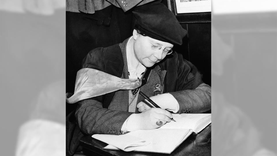 Dorothy L. Sayers, in glasses and beret, signs an open visitors' book with a quill pen.