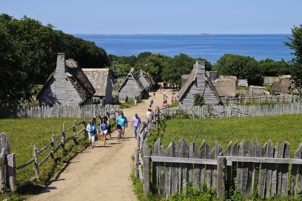 People walking along the walkway from Plimoth Patuxet Museum, Plymouth, Massachusetts, during summer, old wooden buildings with a matching fence, surrounded by trees and ocean in the background