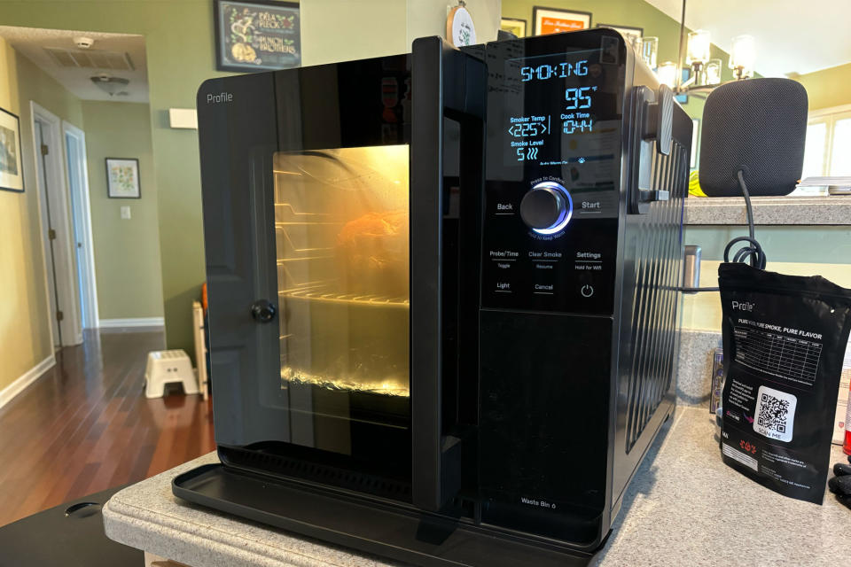 <p>GE Profile Smart Indoor Smoker from the front right corner. Smoker is running, cooking a pork butt inside with the interior light on.</p>
