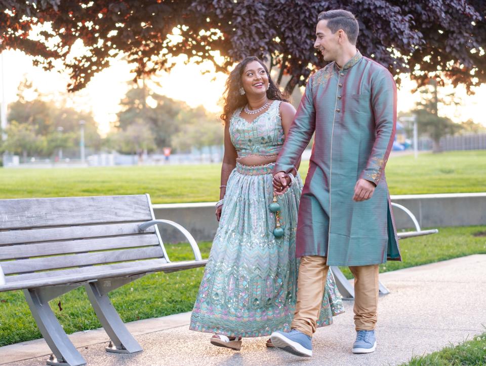 The couple are in matching teal blue traditional Indian clothing walking hand-in-had through a park.