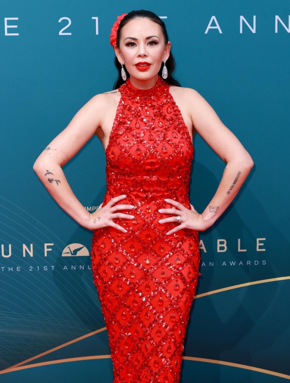 Janel Parrish in an embellished red gown with hands on hips, standing against event backdrop