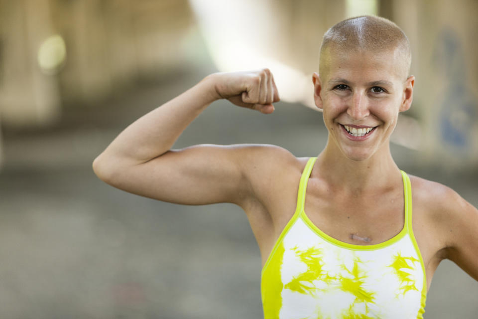 Person with a shaved head flexing their arm muscle, smiling at the camera, wearing a tank top