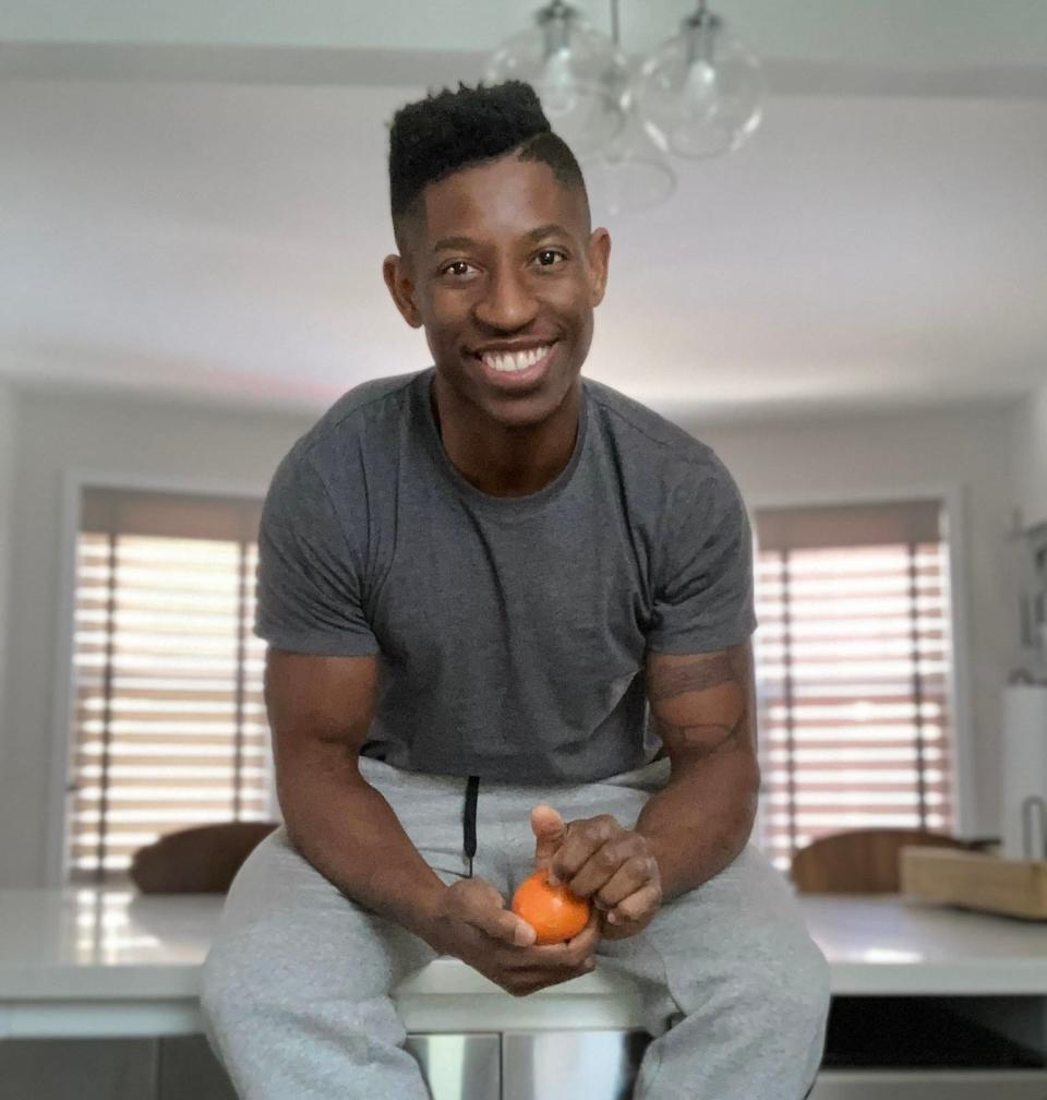 Man seated inside home, smiling at camera, holding an orange. He wears a T-shirt and jogging pants