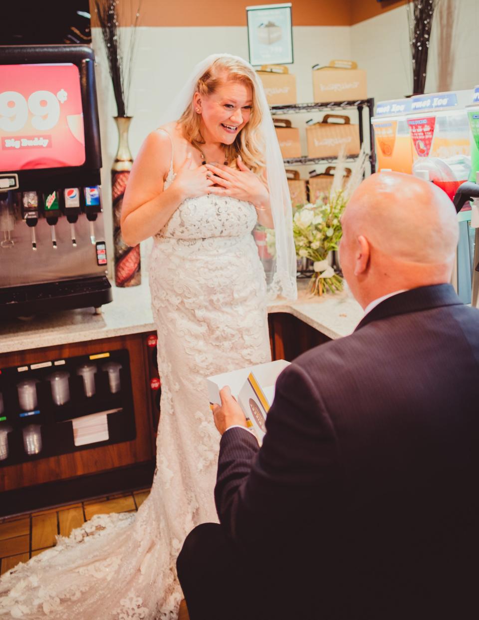 A groom kneels in front of a bride at a gas station.