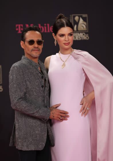 Marc Anthony in a gray suit posing with his hand to belly of his wife Nadia Ferreira who is wearing a light pink dress