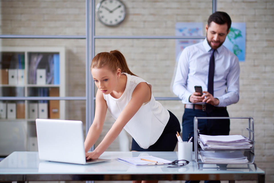 Woman hunching over a laptop at a desk as a man in a tie and shirt stands behind her looking down at his cellphone