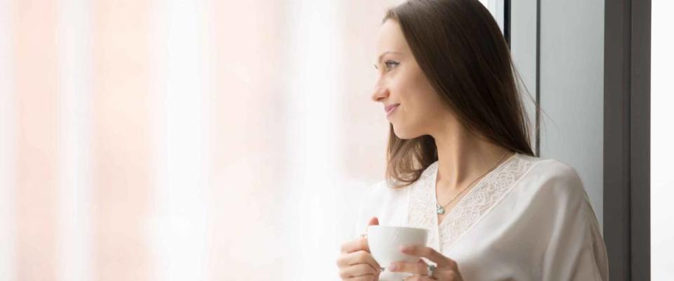 Young smiling woman in lace night gown relaxing at cozy home, feeling happy, resting in the morning, looking through the window with a cup of drink, challenge of a new day. Copy space