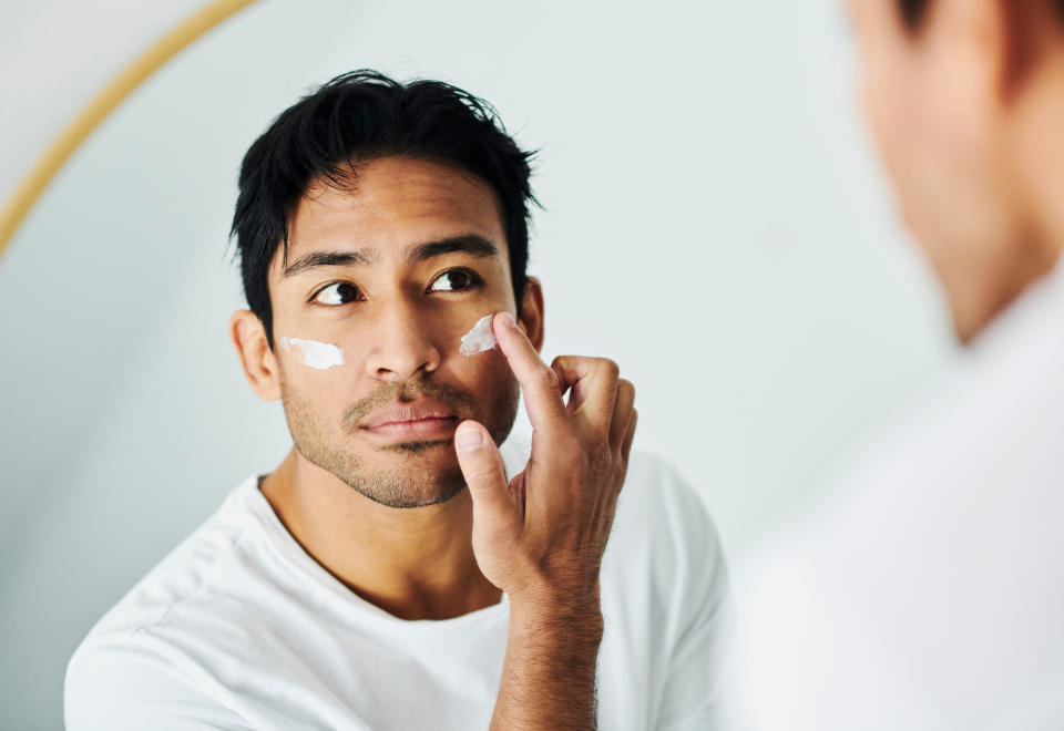 A man in a white shirt applies moisturizing cream to his face while looking into a mirror