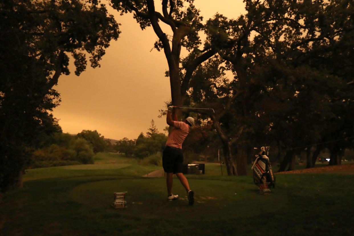 Trees on the green frame an orange sky with a golfer teeing off. 