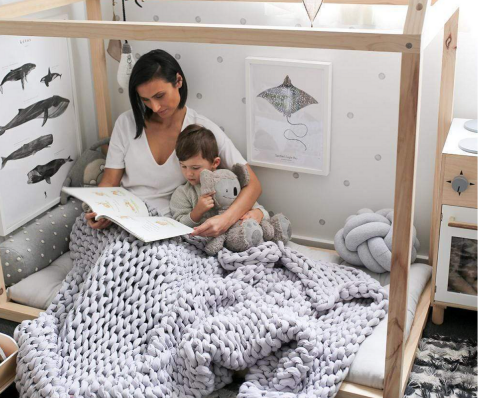 A woman sits snuggled in bed with a child reading a book under a mauve Calming Blanket. 