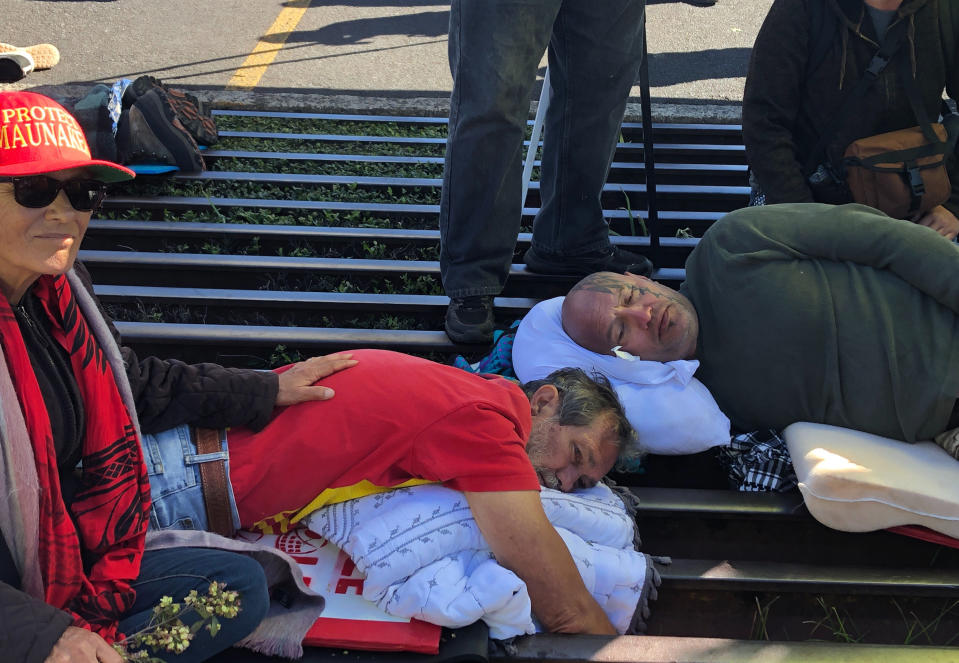 Activist Walter Ritte, left, and others lay chained to a cattle grate blocking a road at the base of Hawaii's tallest mountain, Monday, July 15, 2019, in Mauna Kea, Hawaii, protesting the construction of a giant telescope on land that some Native Hawaiians consider sacred. (AP Photo/Caleb Jones)