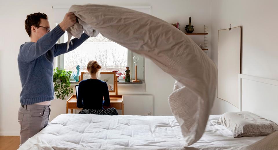 Man makes bed while woman sits at table in background. (Getty Images)