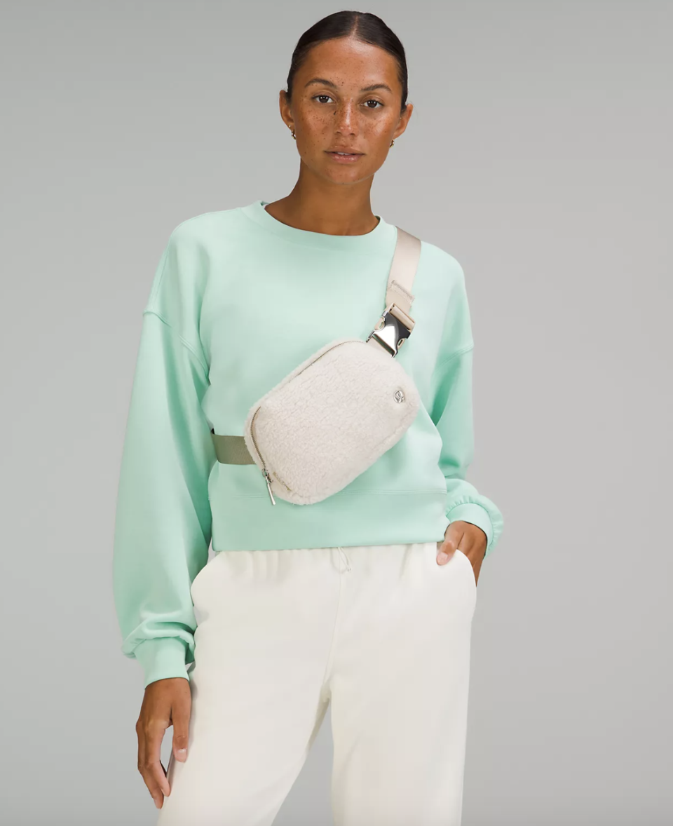 woman wearing white pants, green sweater and Everywhere Belt Bag 1L Fleece in natural ivory (photo via Lululemon)