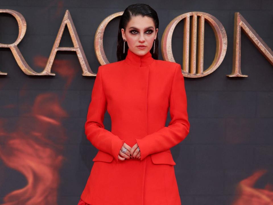 A young woman with dark hair and eye makeup wearing a long red dress and black platform heels.