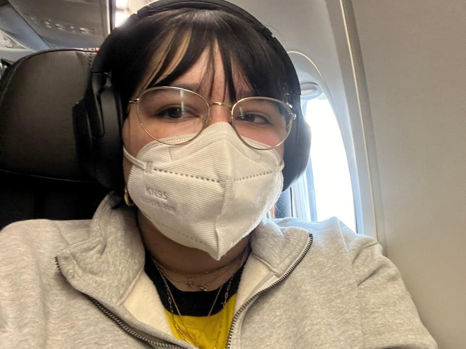 Author Libby Torres sits in a window seat of an American Airlines flight looking at the camera, wearing a gray sweatshirt, yellow t-shirt, black headphones, white KN95 mask, and gold wire-rimmed glasses.