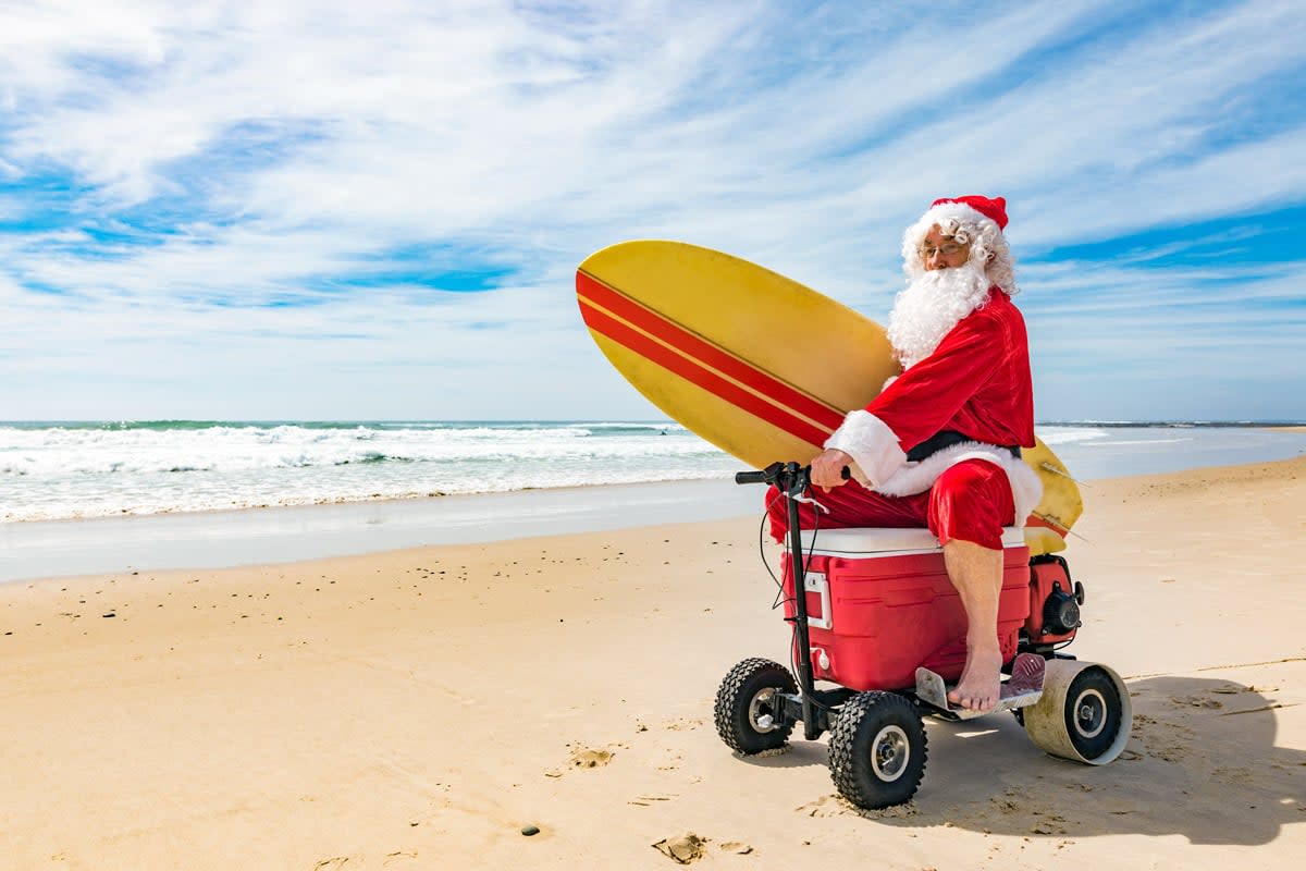 Santa Claus ride a scooter on the beach while holding a surf board. 