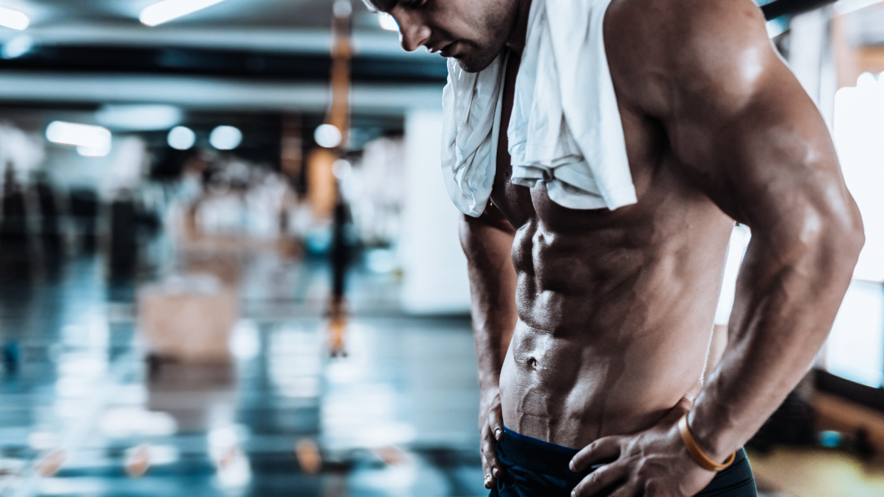  Man standing in a gym topless with a towel round his neck. 