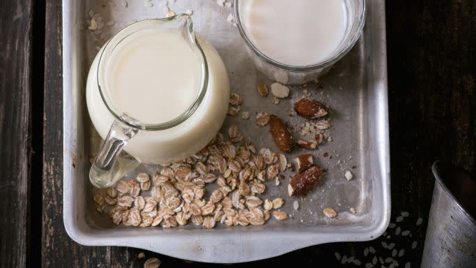set of non dairy milk rice milk, almond milk and oat milk in glass cups and jug on old aluminum tray with rice grains, oat flakes and almond over old wooden table dark rustic style top view
