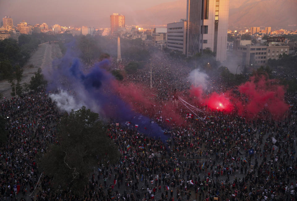 Clouds of smoke from flares hover over a group of anti-government protesters gathered in the Plaza Italia, in Santiago, Chile, Friday, Nov. 8, 2019. Chile's unrest began last month over a subway fare hike. But it has morphed into a movement demanding a broad range of changes. (AP Photo/Esteban Felix)