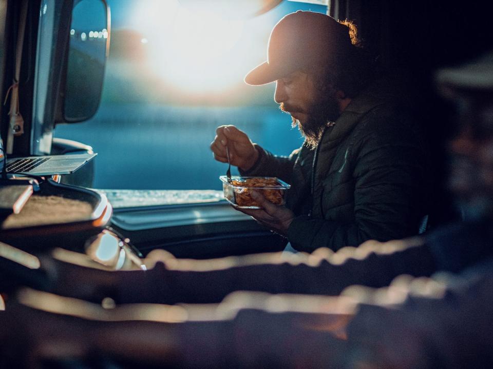 Close up of a trucker driving while his trucking partner is using a laptop and eating