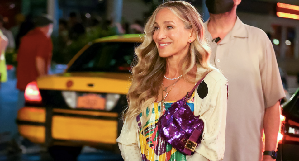 Sarah Jessica Parker smiles as she wears a bright purple sequinned handbag over her shoulder. Her long blonde streaked hair is worn curly and loose. A man wears a mask behind her and a yellow car is parked behind.