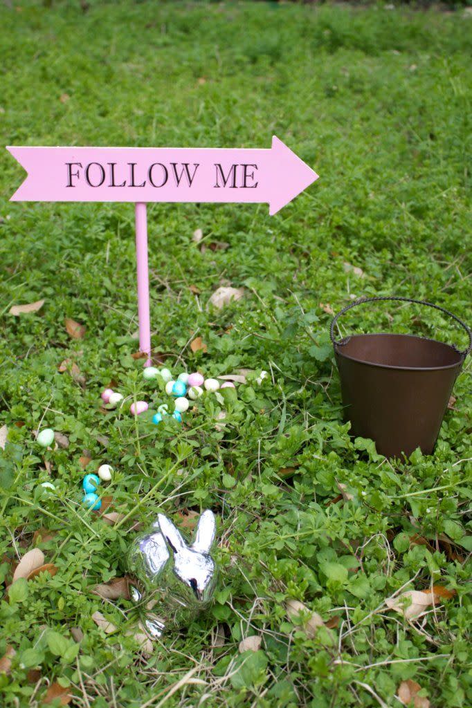 easter egg hunt arrow sign that says follow me, next to candy eggs scattered in grass, silver bunny statue, and metal bucket