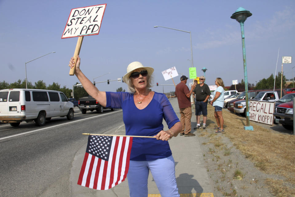 Katherine Hayes waves a flag and a sign urging Alaska lawmakers to fund a full oil wealth fund check, known locally as the PFD or Permanent Fund Dividend, Monday, July 8, 2019, in Wasilla, Alaska. Some Alaska lawmakers are meeting in Wasilla July 8 instead of Juneau, where state House and Senate leadership have decided to hold the special session, called to determine the amount of this year's check. (AP Photo/Mark Thiessen)