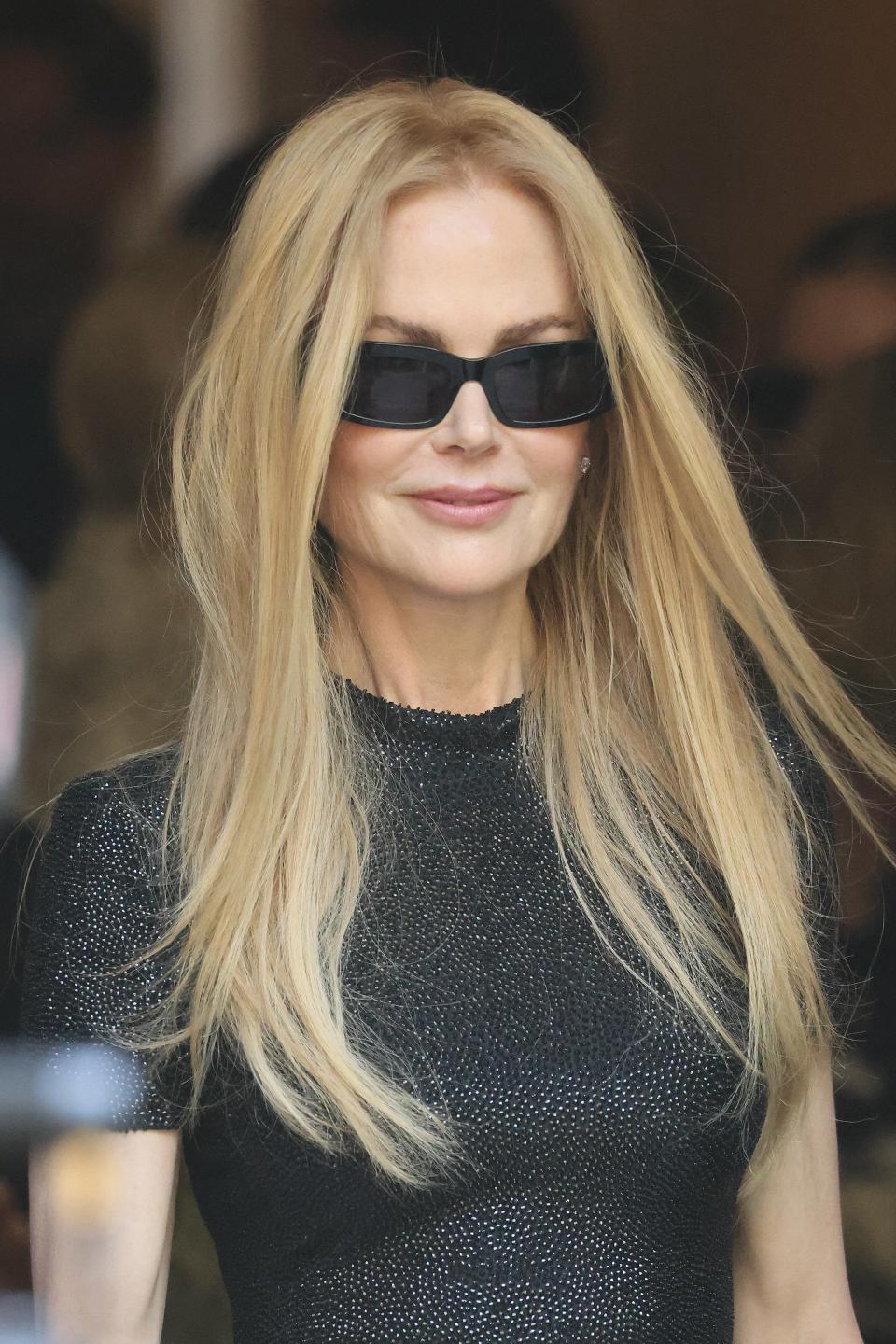 Nicole Kidman, wearing black sunglasses and a fitted black dress, smiles at an event