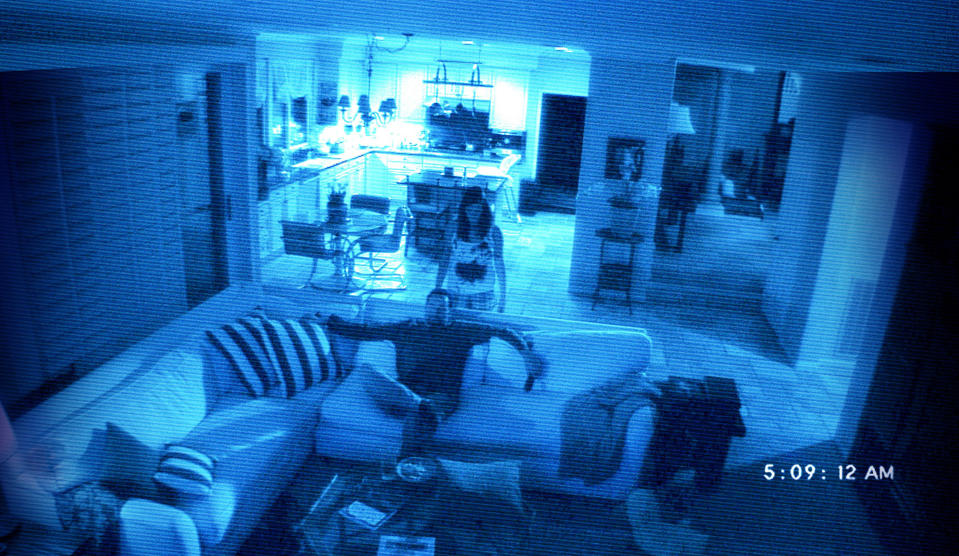 Katie Featherston as Katie stands behind a couch, covered in blood, as Brian Boland playing Daniel Rey sits watching TV in Paranormal Activity 2