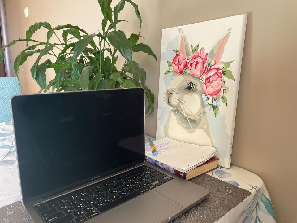 A MacBook with a black screen sits on a table in front of a plant and a painting of a bunny with a flower crown