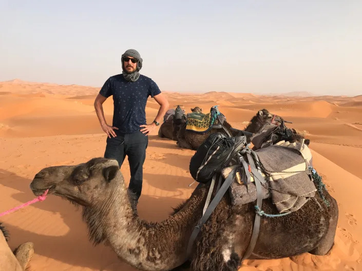 A man standing with hands on hips next to a camel in the desert.