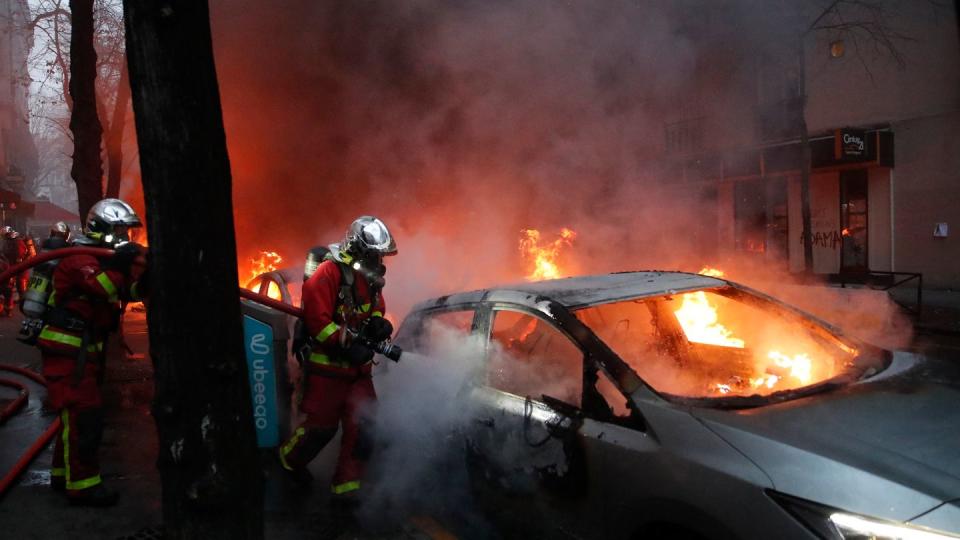 Feuerwehrleute löschen während eines Protestes auf einer Straße ein brennendes Auto.