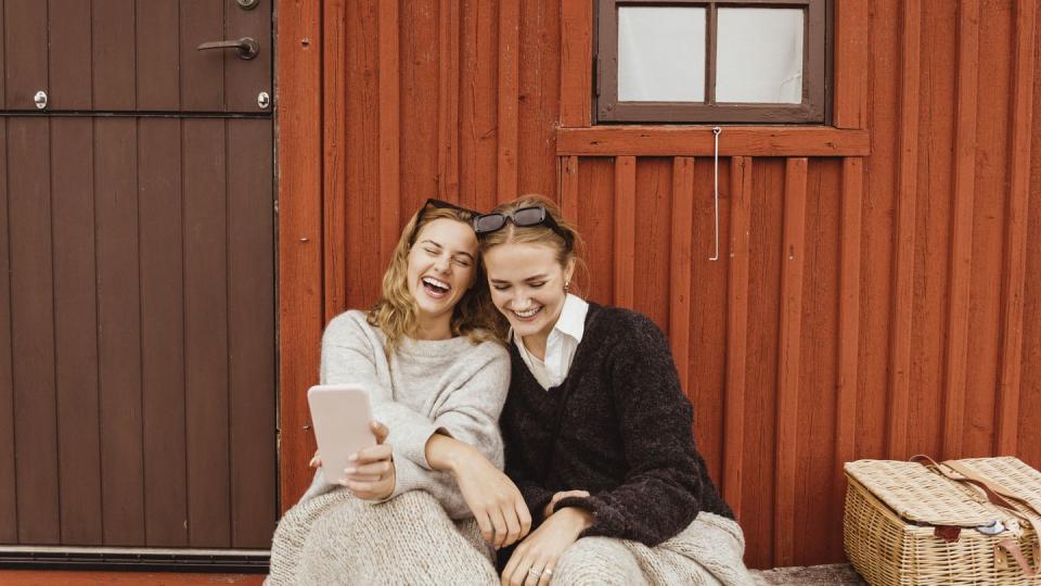 female friends laughing while taking selfie on mobile phone against cottage