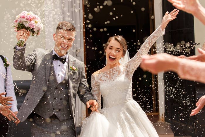 Bride and groom exit wedding venue smiling and holding hands with guests throwing confetti