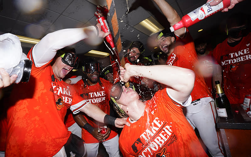 Baltimore Orioles' Heston Kjerstad drinks from the "Homer Hose" while being sprayed by teammates during a locker room celebration. The man in the center is crouched slightly, leaning back to drink while others splash him from bottles of Budweiser. They are all wearing goggles and bright orange T-shirts that read "Take October."