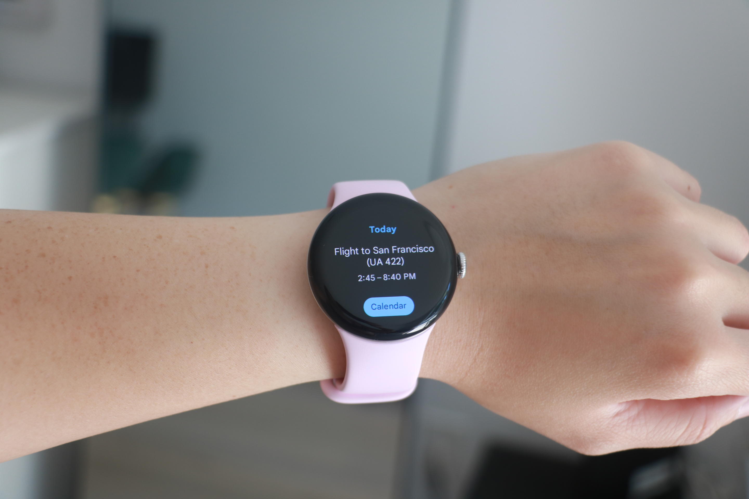 The Pixel Watch 3 with a pink band on a person's wrist in front of a mirror. Its screen shows the word 