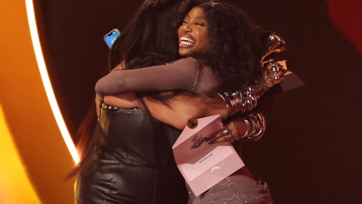 lizzo and sza hug as sza smiles widely, both wear dresses, and lizzo holds a grammy trophy that sza won