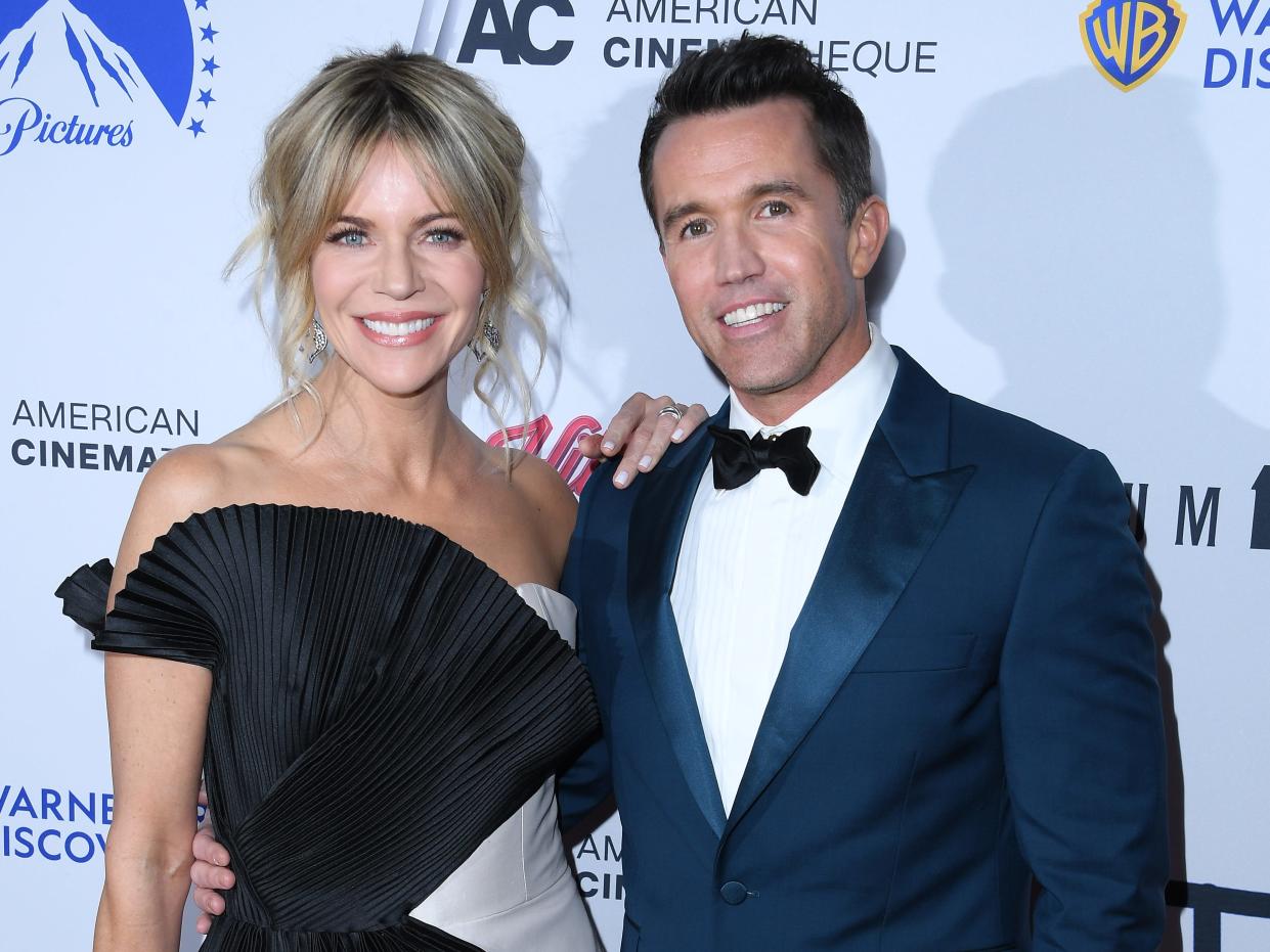 Kaitlin Olson, who is wearing a blade and gray dress, and Rob McElhenney, who is wearing a blue suit with a bowtie, posing for photographers together while attending a press event.