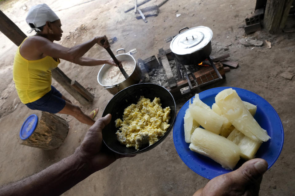 A Indigenous Tembe woman prepares food on a stove in the kitchen at the back of her house, as another resident carries dishes with scrambled eggs, left, and cooked manioc, in the Tenetehar Wa Tembe village in the Alto Rio Guama Indigenous territory, in Paragominas municipality, in Para state, Brazil, Saturday, June 10, 2023. (AP Photo/Eraldo Peres)