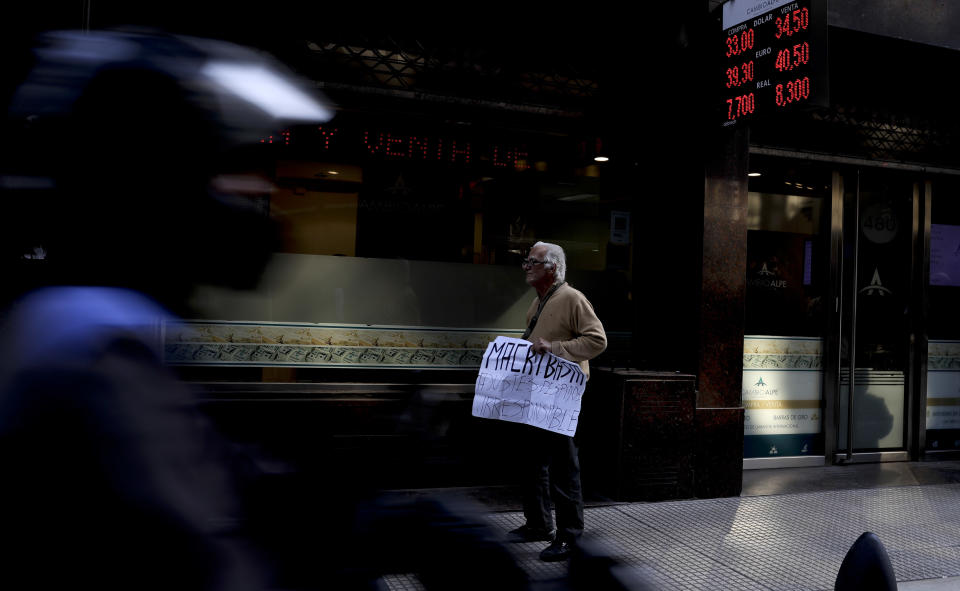 FILE - In this Aug. 29, 2018 file photo, a man holds a sign that says in Spanish "Macri enough adjustments. Firings. Irresponsible." outside a money exchange office in Buenos Aires, Argentina. In 2018 the Argentine peso lost more than half its value to the U.S. dollar, and now Argentines are losing purchasing power to a nearly 50% annual inflation rate, one of the world’s worst. (AP Photo/Natacha Pisarenko, File)