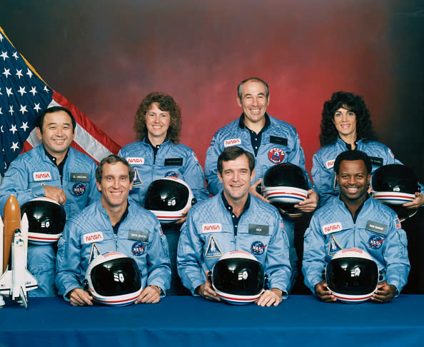 Group photo of seven NASA astronauts in blue uniforms holding helmets. L-R: Ellison Onizuka, Judith Resnik, Ronald McNair, Mike Smith, Dick Scobee, Christa McAuliffe, Gregory Jarvis