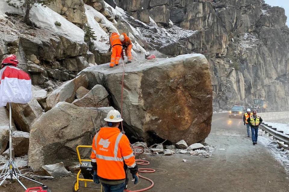 In this photo provided by Caltrans, a crew drills holes to place explosives into a large boulder that fell onto U.S. Highway 50 near South Lake Tahoe, Calif., Friday, March 4, 2022. The return of winter weather prompted forecasters to advise people planning mountain travel to be prepared for snowy roads Friday and Saturday. Travel through the Sierra was disrupted by the massive boulder that fell onto the highway at Echo Summit on Thursday. (Caltrans via AP)